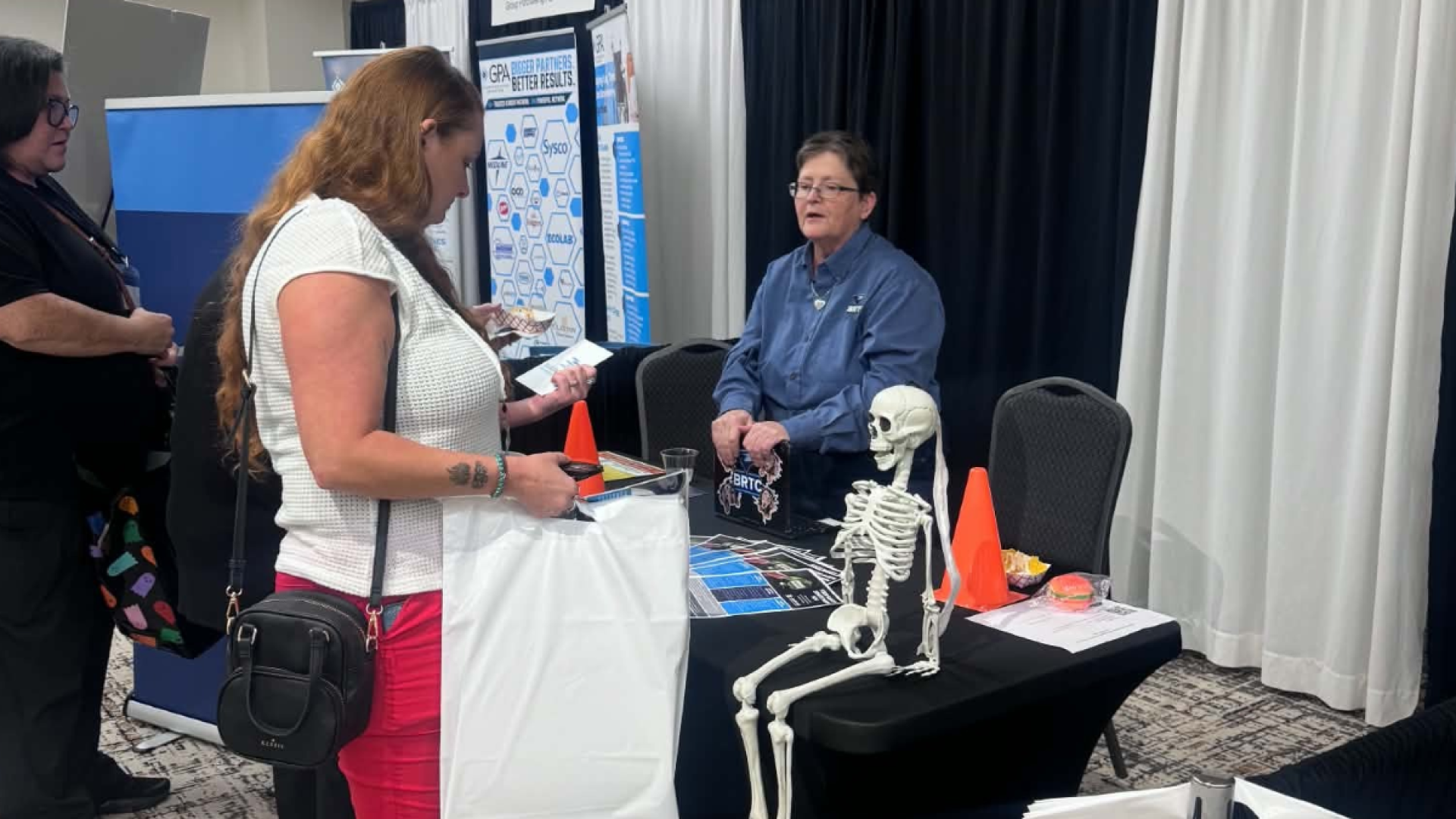 A BRTC representative sits at a conference booth decorated with a skeleton decoration and traffic cones, speaking with an attendee browsing informational materials.